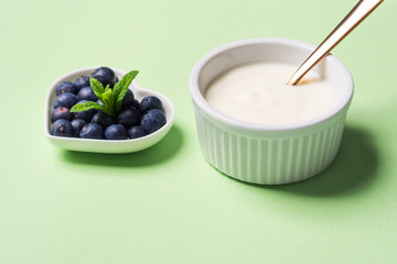 Bowl of natural yogurt and fresh blueberries in heart shaped plate on pastel green background. Healthy breakfast concept