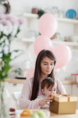 Beautiful young mother holding baby daughter on lap, mom and girl opening birthday gift box together. Room decorated with flowers and pink balloons