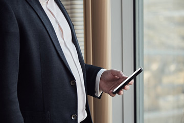 Close up of young businessman in black suit using smart phone while staying at his office.