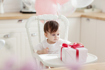 Fototapeta na wymiar Beautiful baby girl sitting in highchair, eating biscuit and looking at gift box with red ribbon on her birthday