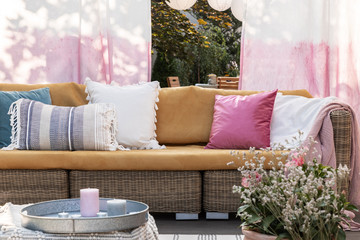 Colorful pillows on rattan sofa on the terrace with flowers, painted drapes and candles. Real photo