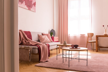 Pink blanket on sofa next to tables on purple carpet in flat interior with drapes at window. Real photo
