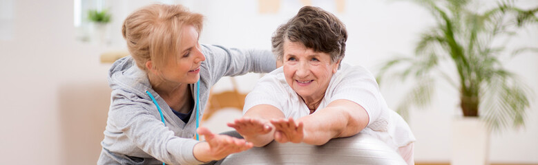 Panorama capture of pilates instructor helping senior woman to stretch on the exercising ball