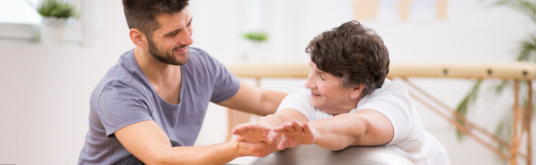 Panorama photo of young physiotherapist helping old paralyzed woman in exercising