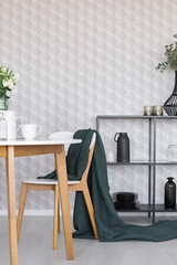 Dark green blanket on wooden chair next to table in stylish dining room interior with metal shelf with plates and jugs and geometric pattern on wallpaper