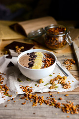 asty breakfast from chocolate smoothie bowl topping with banana, homemade granola and chocolate pieces with coconut milk served on the wood table early in the morning