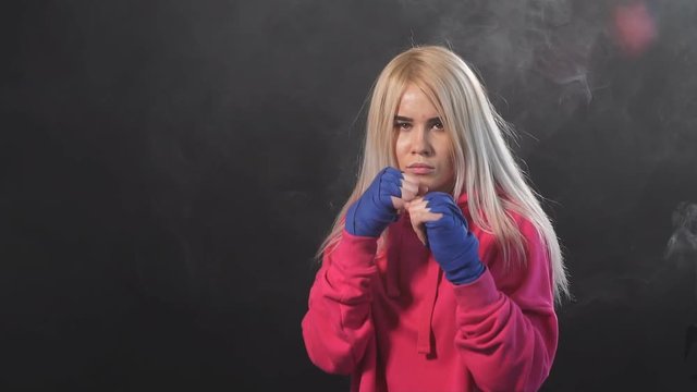 Blonde female athlete is exercising in defense and attacks on dark smoky background, slow motion