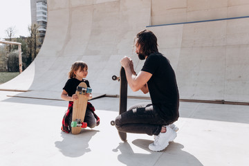 Father and his son dressed in the casual clothes are siting next to the skateboards in a skate park at the sunny day