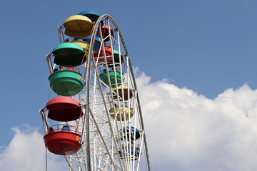 Ferris wheel with colourful cabins against blue sky and white clouds. People ride the damn wheel in amusement park