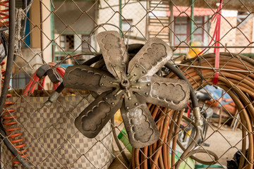 Rusty Old Fan Blade/ Clutch Hanging on Metal Chain-link Fence - Vintage Messy Garage with Dirty Plastic Hose and PVC Pipe