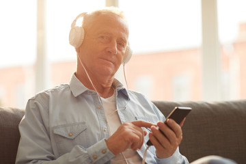 Portrait of modern senior man wearing headphones using smartphone sitting on sofa at home and looking at camera