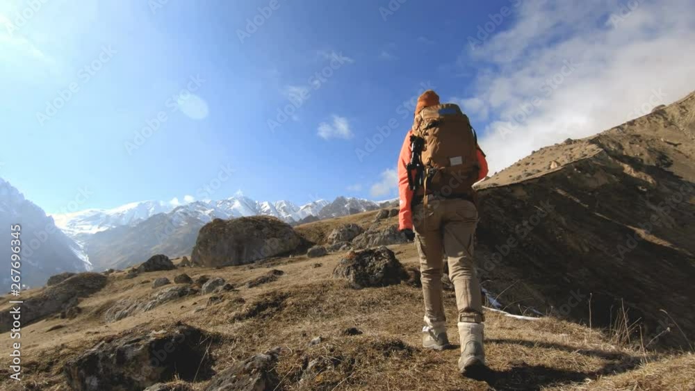 Wall mural Happy traveler girl photographer in sunglasses with a camera and a backpack goes uphill on a background of snow-capped peaks on a sunny day