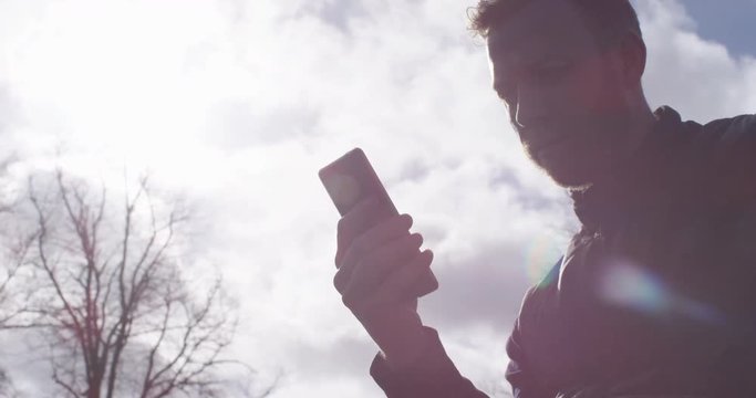 4K Silhouette of man with smartphone against background of cloudy sky & trees. Slow motion.