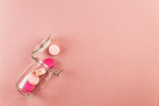 Pink french macarons or macaroons, falling out of a glass jar over a pink tablecloth background with copy space.