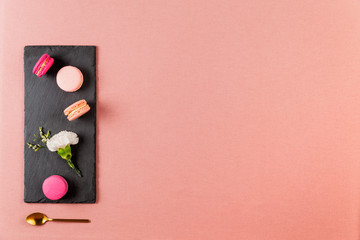 French pink macarons or macaroons, and incarnation white flower on a slate, with golden tea spoon over a pink tablecloth background with copyspace.