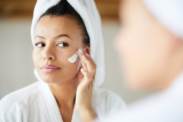 Head and shoulders portrait of  beautiful Mixed-Race woman applying face cream during morning routine, copy space - obrazy, fototapety, plakaty