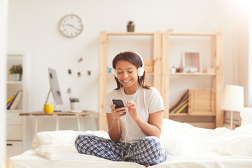 Portrait of young Mixed-Race woman using smartphone sitting on bed lit by sunlight, copy space