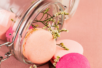 Light pink and fuchsia french macarons or macaroons, falling from a glass jar with light yellow and white tiny flowers over a pink tablecloth,  closeup.