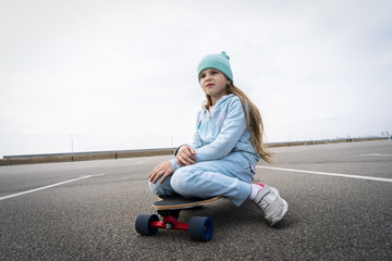 A girl in a blue hat and sweater sits on a large longboard along a large asphalt ground.