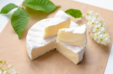 a camembert cheese wheel lying on a table among white flowers and green leaves, two pieces cut off, overhead view