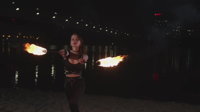 Stylish young woman showing motion of fire juggling burning torches standing on riverbank at night. Skillful female juggler showing mastery of work with flame outdoor, with night city in background.