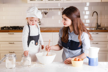 The little daughter in the chef's hat and apron and her mother prepare baking in the bright, classic kitchen.