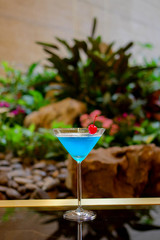 Blue cocktail in martini glass decorated with red cherry and green plants as background