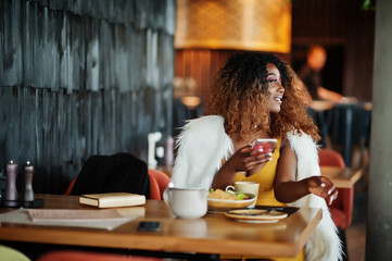 Glamour african american woman in yellow dress sitting at the table with dishes on restaurant with mobile phone at hand.