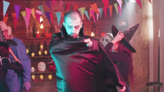 Man in vampire costume hides behing the cloack at a halloween party. Friends having fun disguised in different scary characters in a pub.