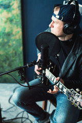 Fototapeta na wymiar Close up of a man singer sitting on a stool in a headphones with a guitar recording a track in a home studio