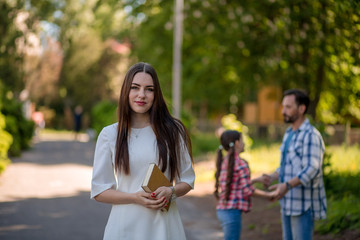 Young Woman Is Holding The Book While Standing In The Park.Her Hasband And Daughter Holding Each Other Hands And Making Circle On The Background.