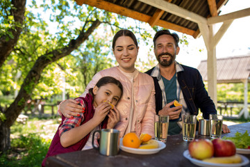 Happy Family Are Sitting By The Table In The Park. Plates With Different Fruits And Glasses With Water Are On The Table.