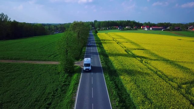 Aerial reveal type clip of a field of bright and yellow rapeseed while two card drive in the highway. Drone static at mediud altitude.