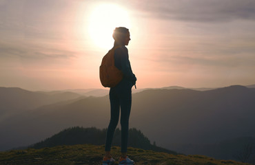 Silhouette of young woman standing on edge of mountain and enjoying life on sunset sky and mountains background. Travel and active lifestyle concept.