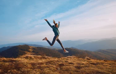 Fotobehang Happy woman hiker jumping on mountain ridge on blue cloudy sky and mountains background. Travel and active lifestyle concept. © vitaliymateha