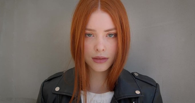 Young female ginger rocker in leather jacket flirting with camera leaning on the white wall.