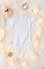 Top view white baby bodysuit on a woolen background. Copy space for text. Accessories and border arrangement with toys and garland of glowing hearts