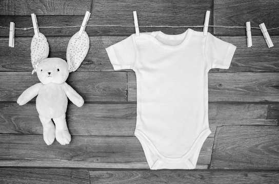 Top view white baby bodysuit on a clothesline with teddy hare on a wooden background. Copy space for text mockup