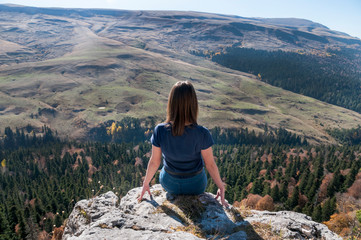Naklejka premium a young active girl sits on the edge of a rock, look at the Caucasus mountains.