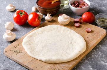 Closeup of raw dough prepared for pizza on the wooden board and different classic ingredients for it on the grey kitchen table