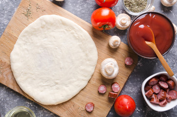 Top view of raw dough for pizza on the cutting board and various ingredients for it.Tomatoes, sauce, mushrooms, oil, herbs, sausages on the grey kitchen table 