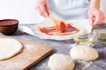 Chef preparing fresh pizza and spreading tomato sauce on dough.Dough balls, bowls with oil, tomato sauce, dry herbs for cooking pizza on the kitchen table