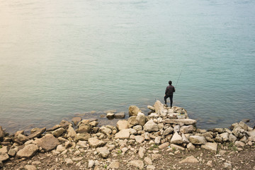 Lonely Asian fisherman standing on rocks in front of water - Powered by Adobe