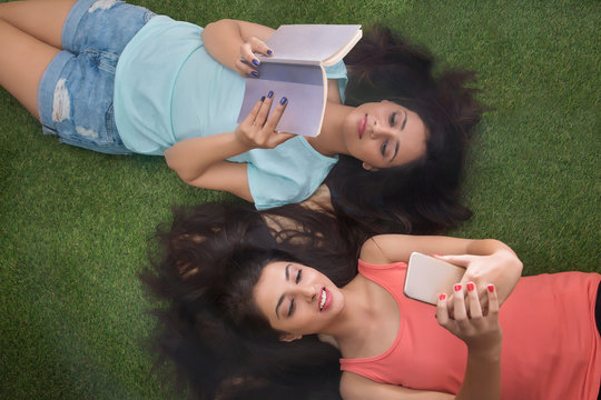 Top view of two teenage girls lying on grass with hair spread all around with one reading book and the other looking at mobile phone	