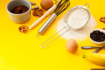 Ingredients for baking with baking utensile on yellow background - flour, wooden spoon, rolling pin, eggs, coccoa, sprinkles, banana. Top view, copy space.