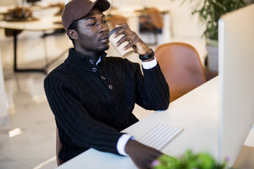 Portrait of young African businessman with cup of coffee looking at pc screen at workplace in modern office