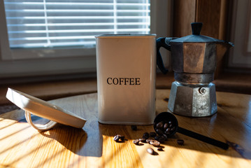 Coffee in a white cup and saucer with silver spoon, a tin of coffee beans, coffee beans and a coffee maker on a kitchen table in bright sunlight