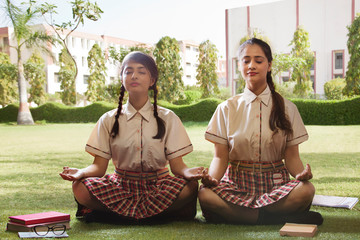 School girls meditating with books kept aside them in the school campus garden	