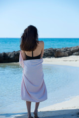 Young girl is on the beach and standing, posing to the camera. Beautiful girl, wrapped in pink white striped beach towel, sunny and windy weather. Background of blue sky, sea and beach - Image