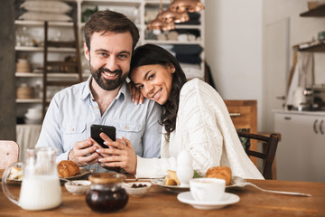 Portrait of adorable couple using mobile phone while sitting at table together during breakfast in kitchen at home